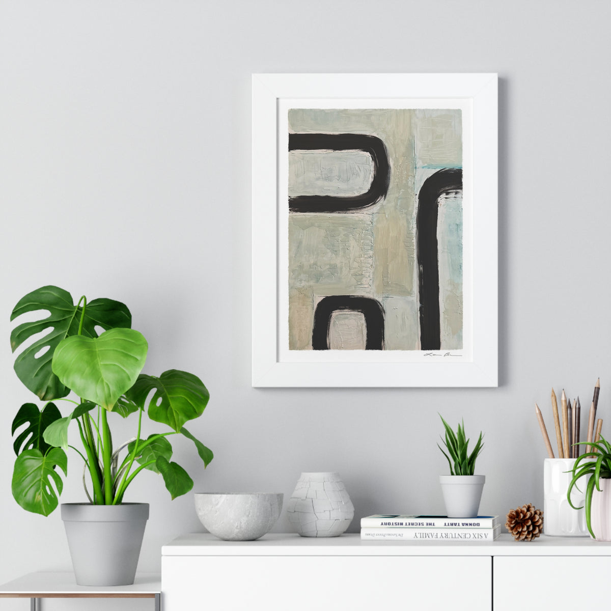 Framed abstract art piece on a wall with plants and decor items below.